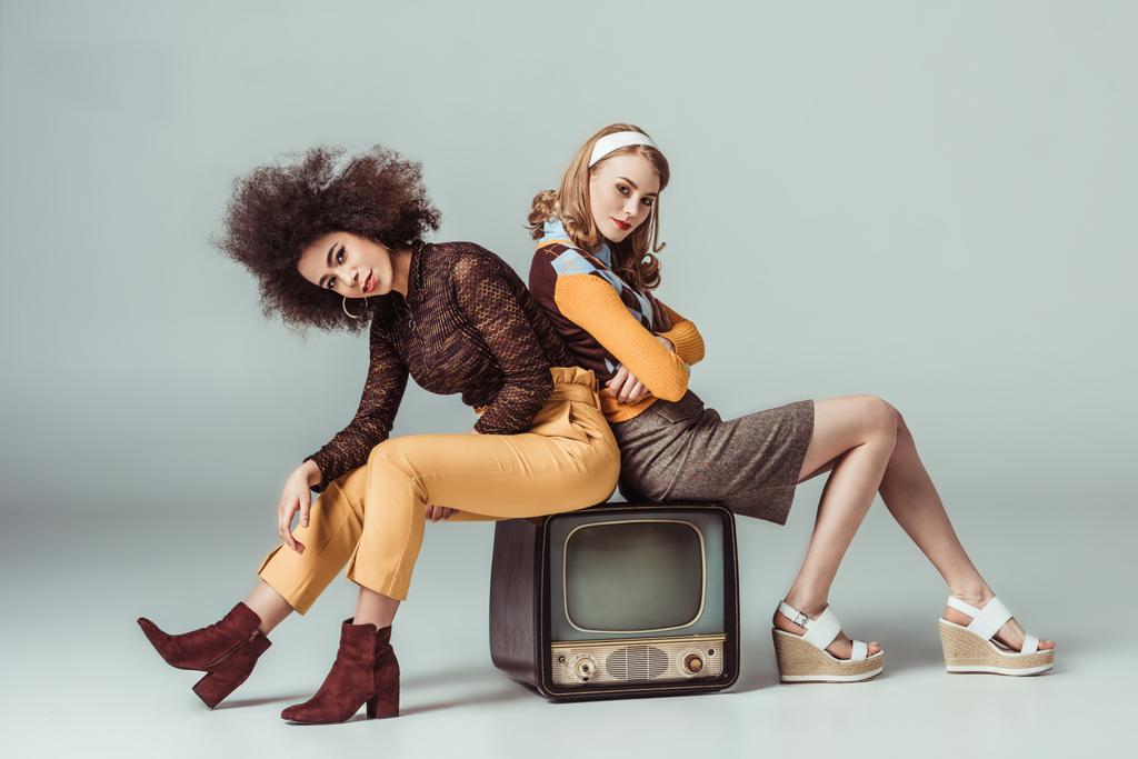 multicultural retro styled girls sitting on vintage television and looking at camera on grey - Photo, Image