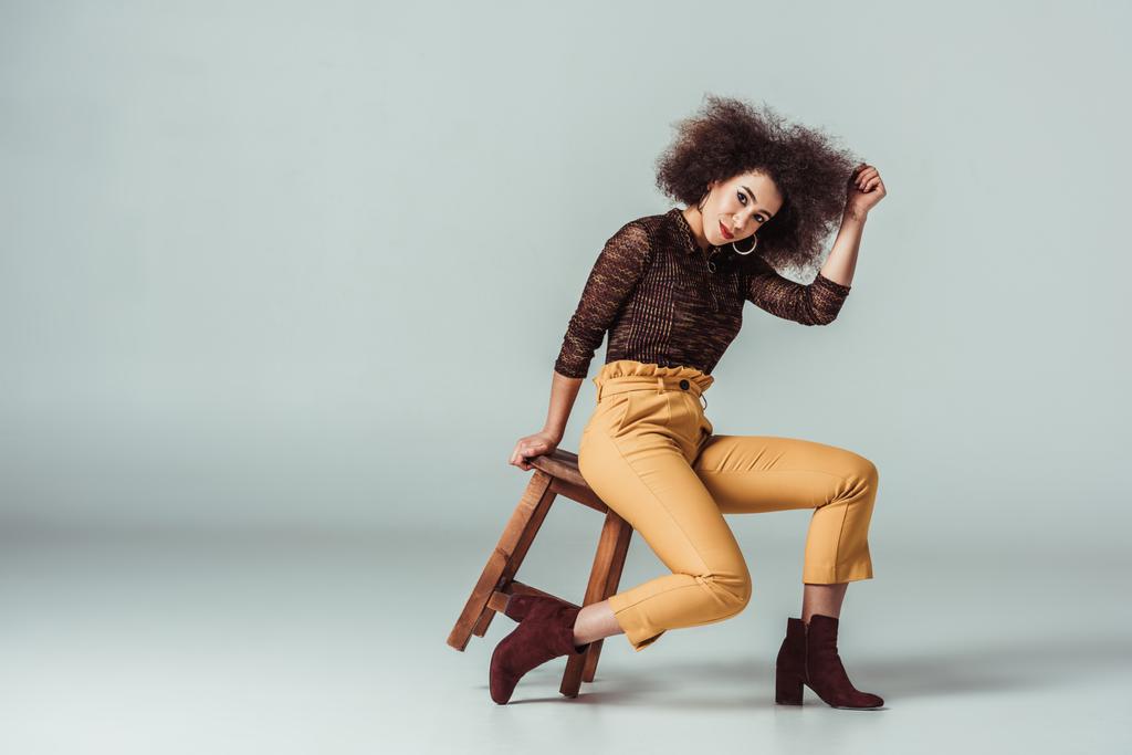 african american woman in retro clothes posing on chair on grey - Photo, Image
