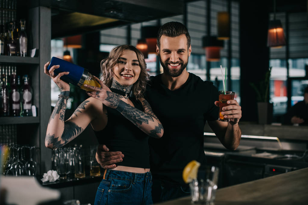 uśmiechający się młodych barmanów przygotowanie napoje alkoholowe w barze i patrząc na kamery - Zdjęcie, obraz