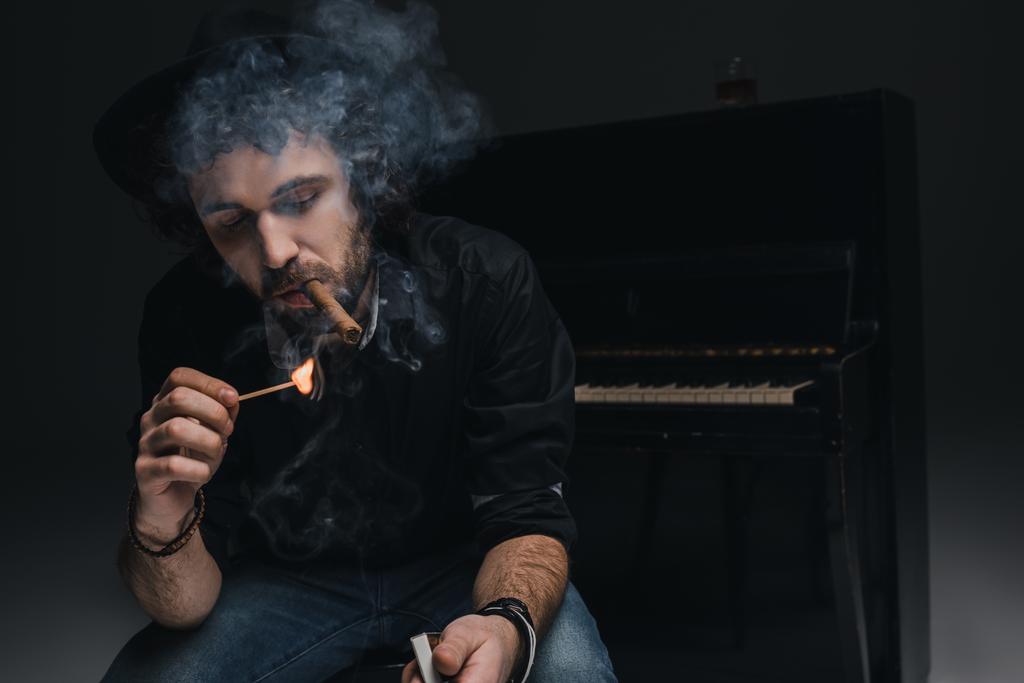 handsome bearded musician smoking cigar in front of piano on black - Photo, Image
