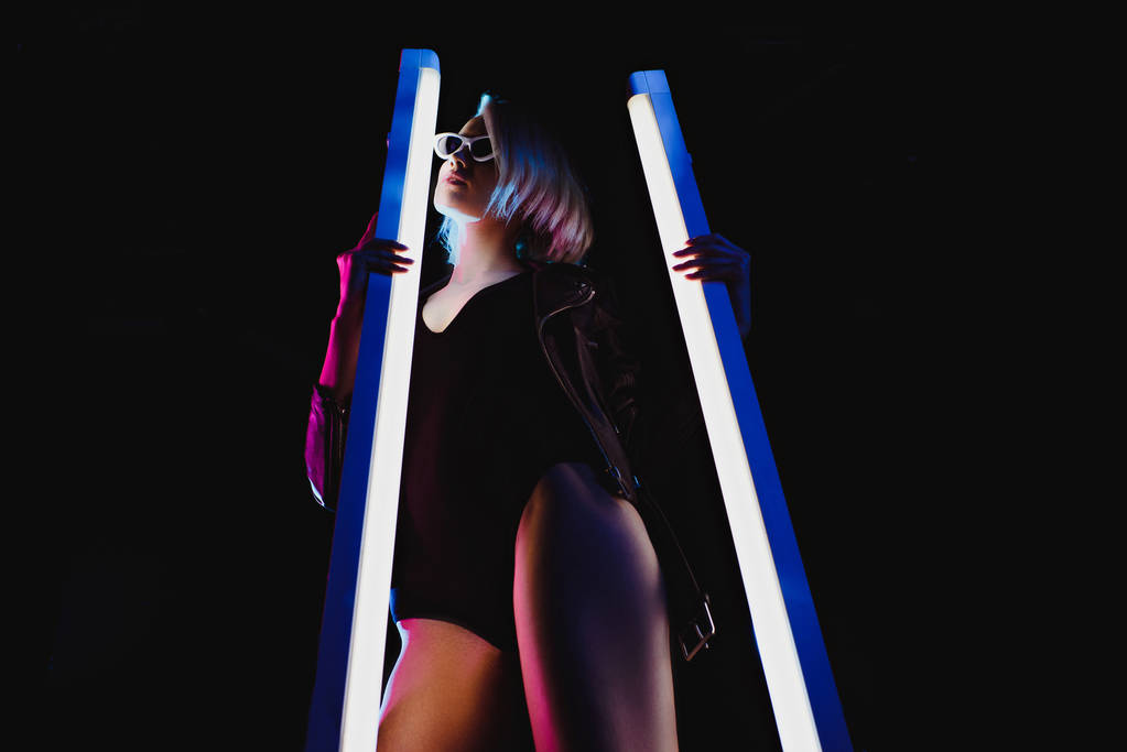 стильная девушка позирует с двумя ультрафиолетовыми лампами для модной съемки, изолированная на черном
 - Фото, изображение