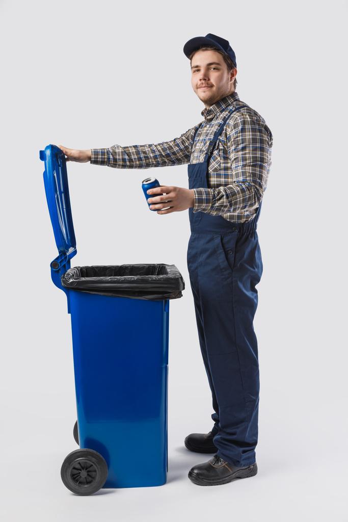 vista laterale di giovane pulitore in uniforme con lattina in piedi al bidone della spazzatura isolato su grigio
 - Foto, immagini