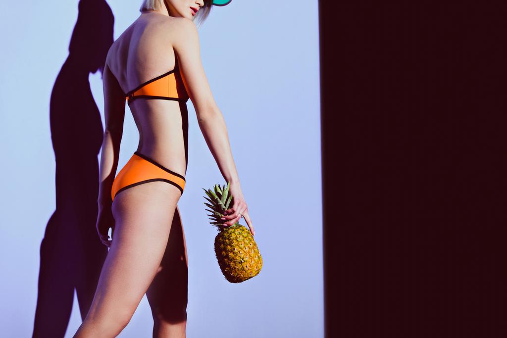 обрезанный вид девушки, позирующей в бикини и солнечном козырьке с ананасом на фиолетовом
 - Фото, изображение