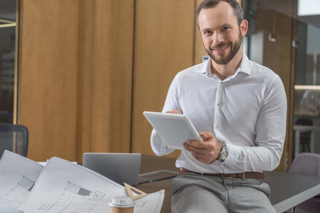 handsome happy architect sitting on table with plans and using tablet at office - Photo, Image