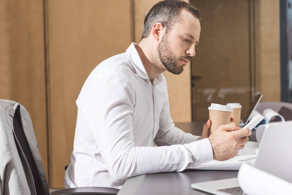 serious architect with paper cup of coffee using smartphone at office - Photo, Image