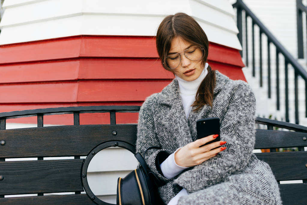 Stilvolles schönes Mädchen in grauem Mantel und Brille blickt in ihr Smartphone, schreibt eine Nachricht, trübes Wetter - Foto, Bild