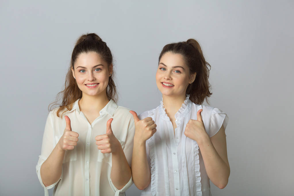 két gyönyörű mosolygó lány nővérek ikrek a fehér blúz, a világos háttér bemutatás gesztusok siker - Fotó, kép