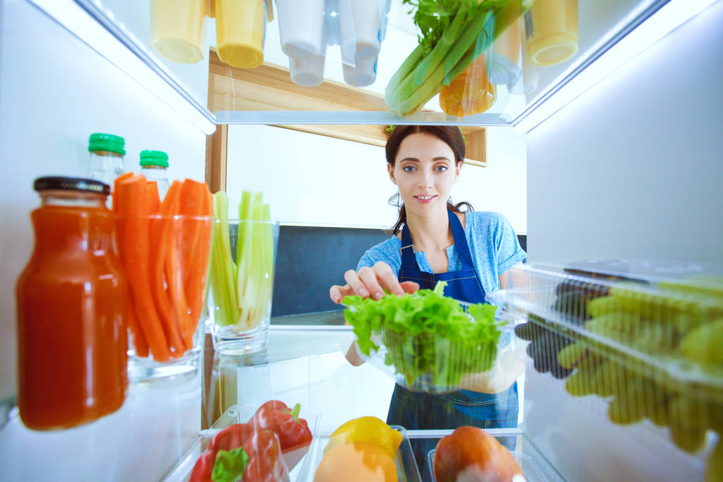 Portret kobiety stojącej w pobliżu otwartej lodówki pełnej zdrowej żywności, warzyw i owoców. Portret kobiety - Zdjęcie, obraz