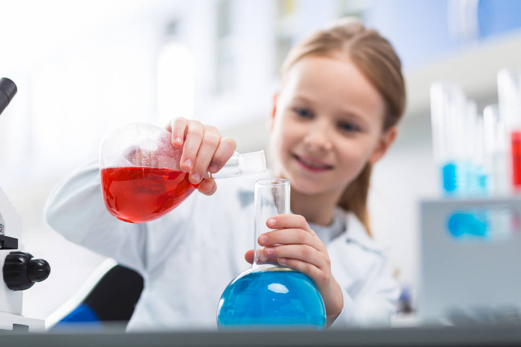 Interesting experiment. Selective focus of glass transparent bulbs hold by little girl who staring at them and dressing in lab coat - Photo, Image