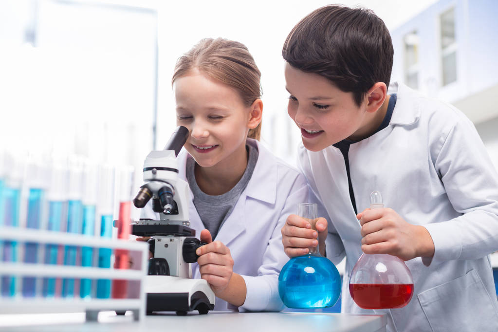 Precise look. Jolly energetic curious girl using microscope and smiling while boy holding bulbs - Photo, Image