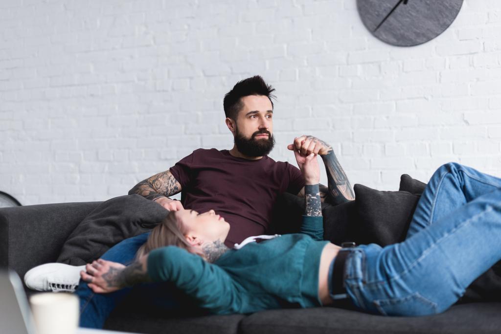 tattooed couple holding hands on sofa in living room - Photo, Image