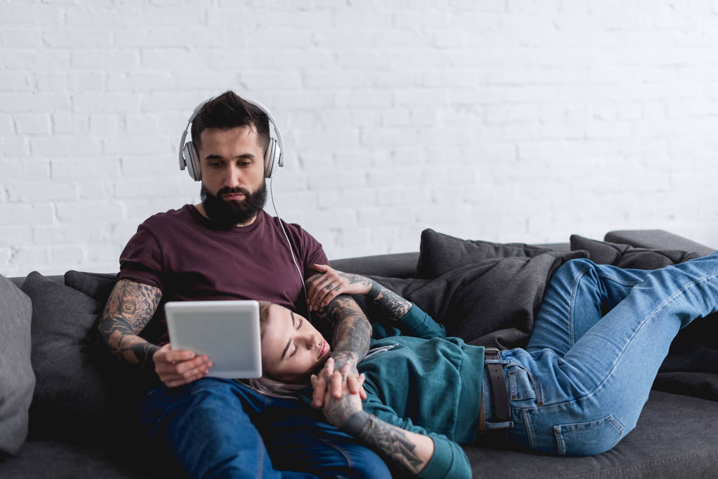 眠っている刺青ガール フレンドと彼氏ソファーにタブレットで音楽を聴く - 写真・画像