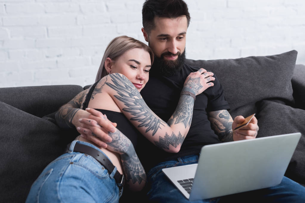 feliz pareja tatuada mirando portátil en sofá en casa
 - Foto, Imagen