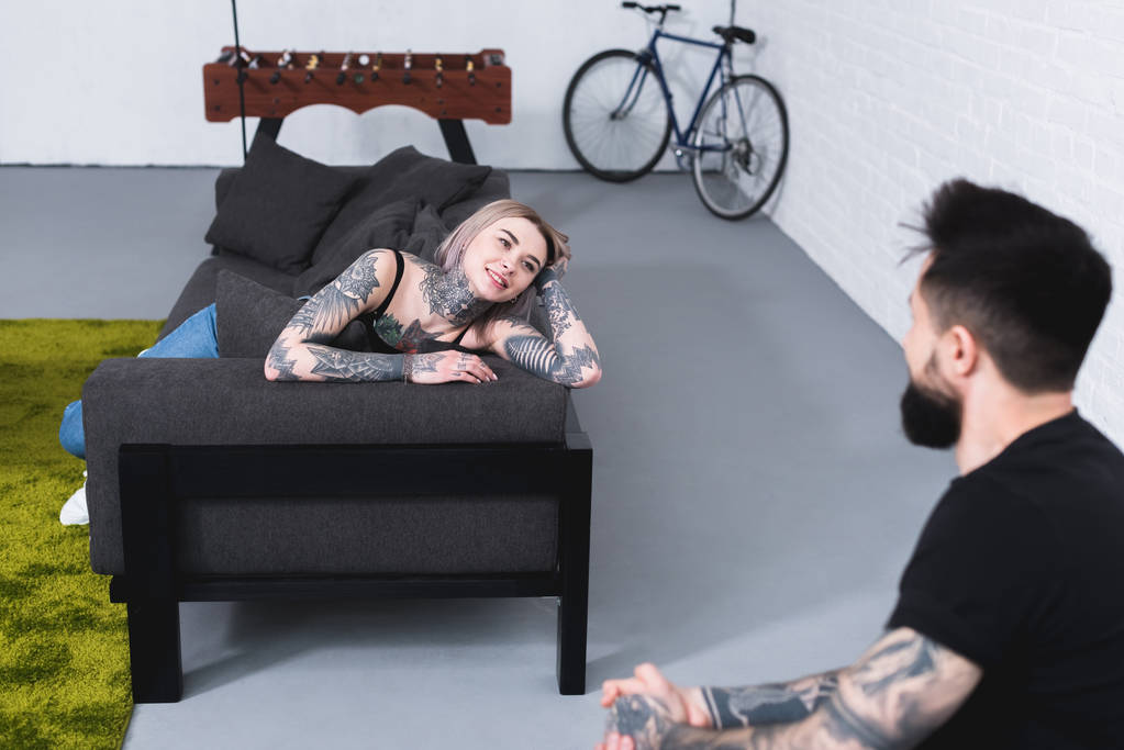 uśmiechający się tatuażem dziewczyna leżąc na kanapie i patrząc na chłopaka w domu - Zdjęcie, obraz