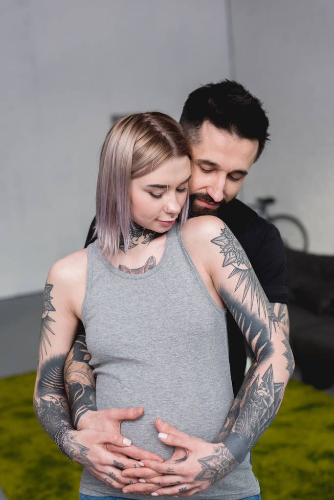 ευτυχισμένος τατουάζ ο φίλος αγκαλιάζει έγκυος κοπέλα από πίσω στο σπίτι - Φωτογραφία, εικόνα