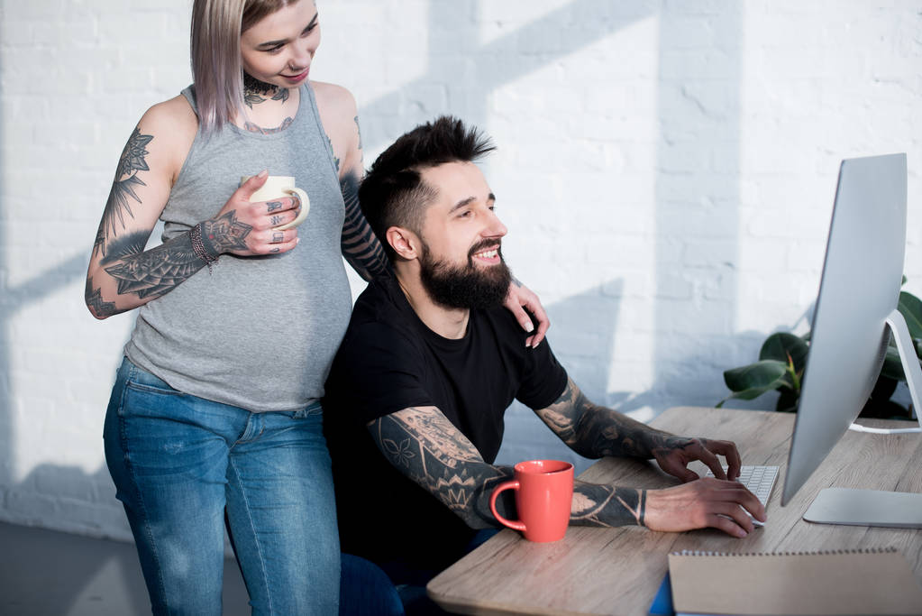 tatuado grávida namorada beber chá, abraçando namorado e olhando para o computador
 - Foto, Imagem
