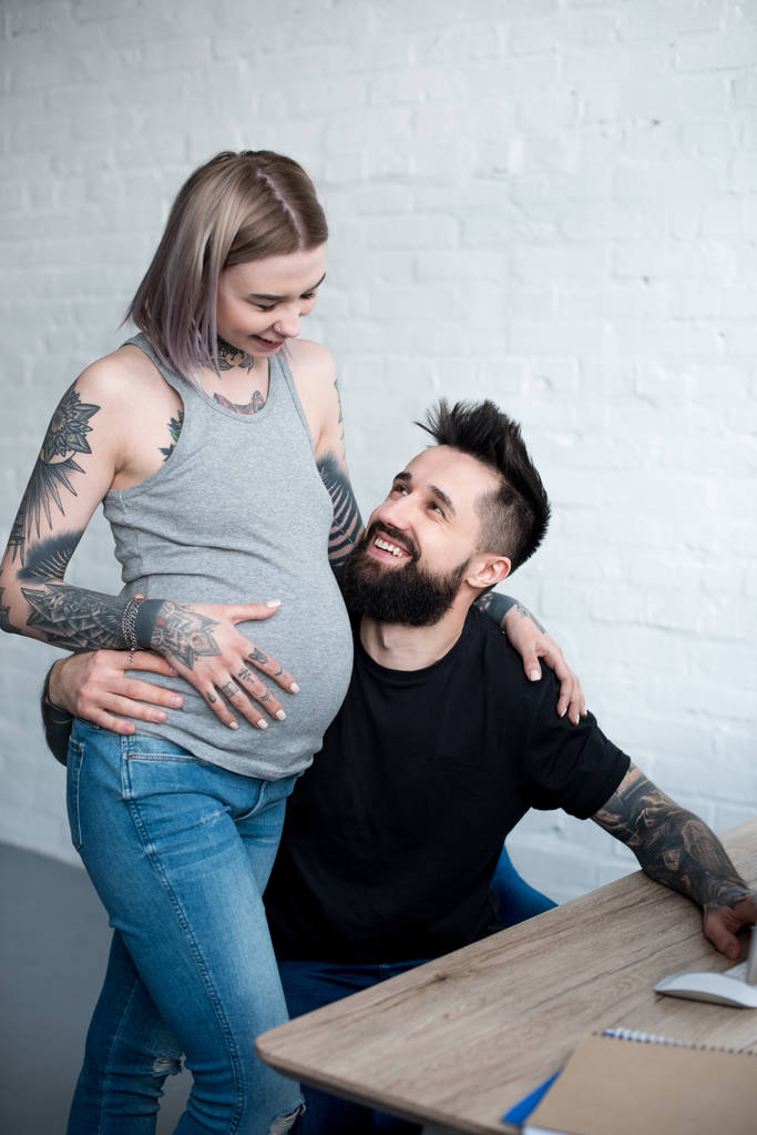 hymyilevä tatuoitu poikaystävä halaa raskaana tyttöystävä kotona
 - Valokuva, kuva