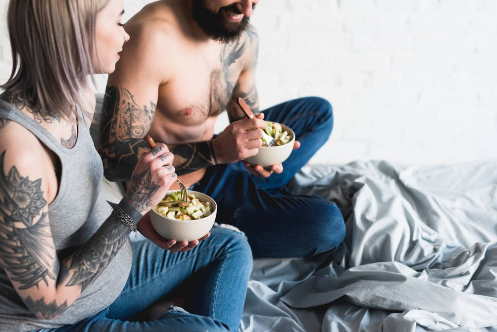 обрезанное изображение беременной татуированной девушки, поедающей салат с парнем без рубашки
 - Фото, изображение