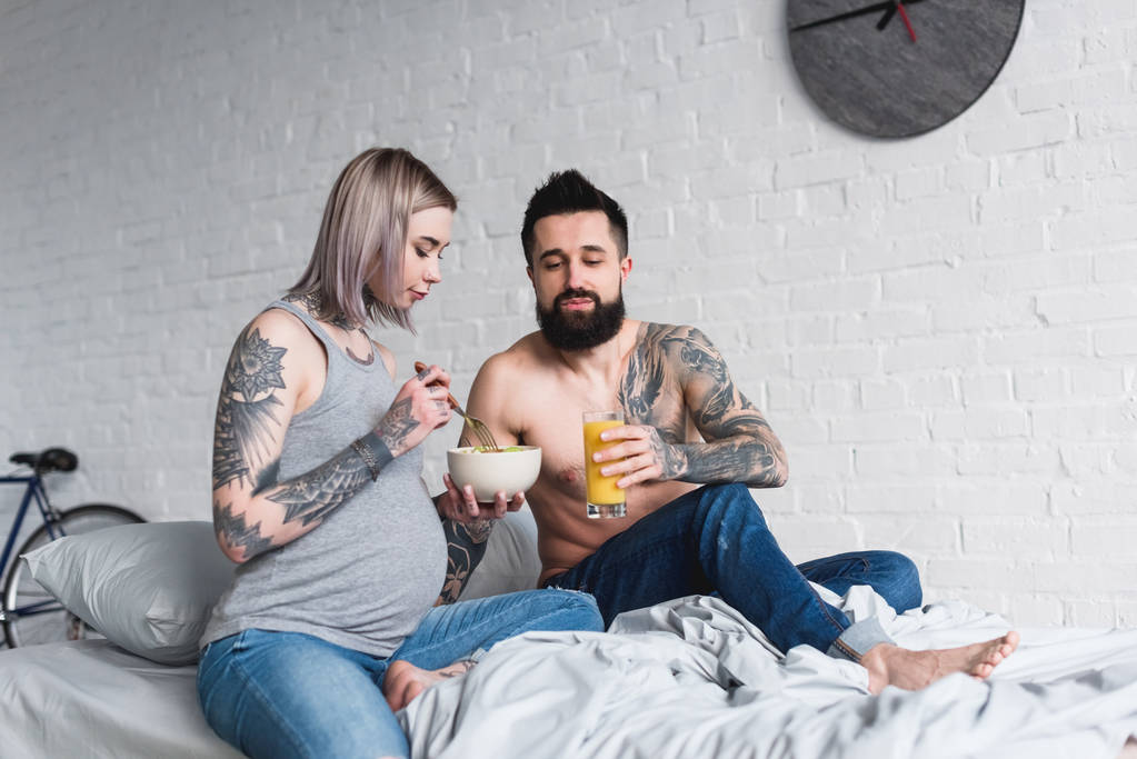 raskaana tatuoitu tyttöystävä syö salaattia sängyssä
 - Valokuva, kuva
