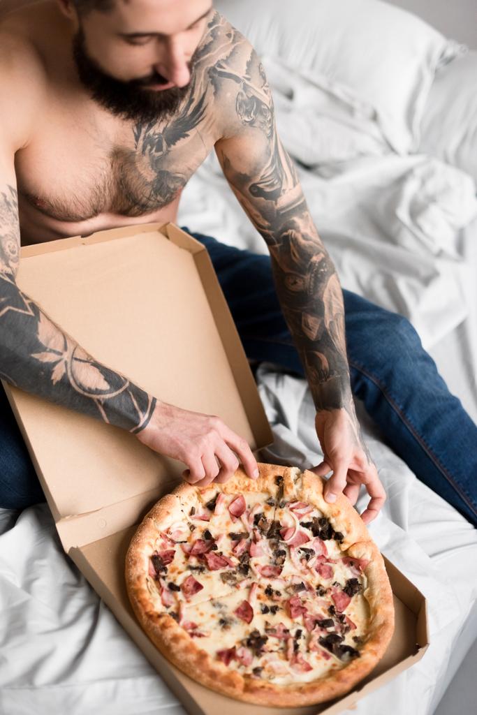 gömleksiz yakışıklı dövmeli adam yatakta pizza ile - Fotoğraf, Görsel