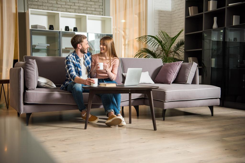 Молода пара сидить на дивані з чашками у вітальні з сучасним дизайном
 - Фото, зображення