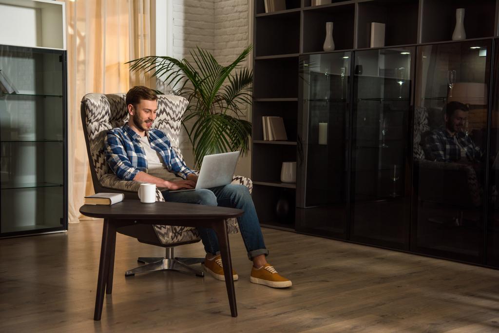 Hombre sentado en sillón y el uso de ordenador portátil en la sala de estar con interior de diseño
 - Foto, imagen