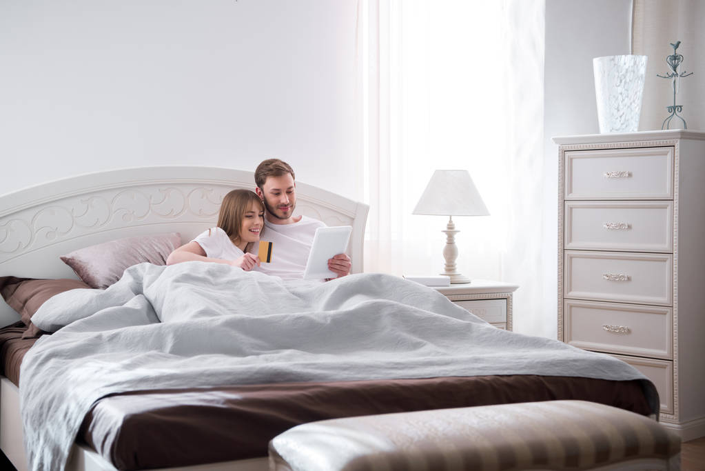 Νεαρό ζευγάρι ξαπλωμένη στο κρεβάτι και να κάνει online αγορές σε μοντέρνα κρεβατοκάμαρα - Φωτογραφία, εικόνα