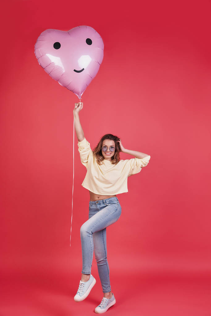 atraktivní mladá usmívající se žena držící srdce ve tvaru balónu a udržet ruku v vlasy stoje růžové pozadí - Fotografie, Obrázek