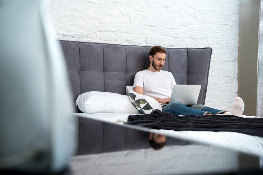 Επιφάνεια επίπεδο ενόψει της νεαρός άνδρας κάθεται στο κρεβάτι με το φορητό υπολογιστή σε σύγχρονη κρεβατοκάμαρα - Φωτογραφία, εικόνα