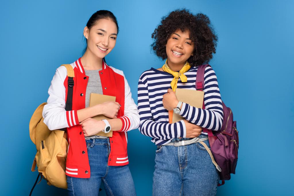 glückliche afrikanisch-amerikanische und asiatische Studentinnen posieren mit Büchern, isoliert auf blau - Foto, Bild