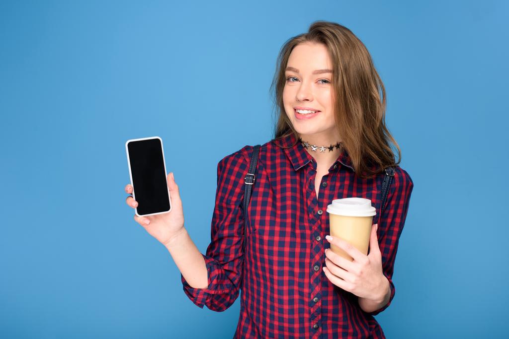 hermosa joven sosteniendo la taza en el café y mostrando teléfono inteligente, aislado en azul
 - Foto, imagen