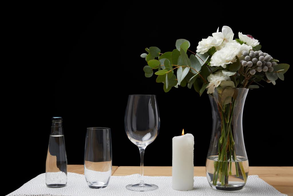 Buquê em vaso com garrafa de água e copos vazios na mesa ao lado da vela no fundo preto
 - Foto, Imagem