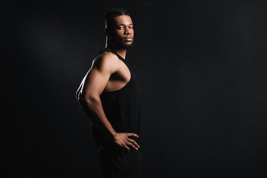 Seitenansicht des athletischen jungen afrikanisch-amerikanischen Mannes, der mit den Händen auf der Taille steht und isoliert auf schwarz in die Kamera schaut - Foto, Bild