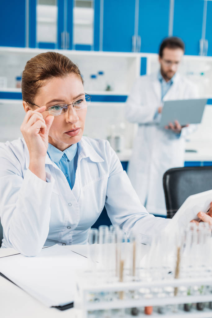 foco seletivo de cientista do sexo feminino usando tablet no local de trabalho com colega atrás no laboratório
 - Foto, Imagem