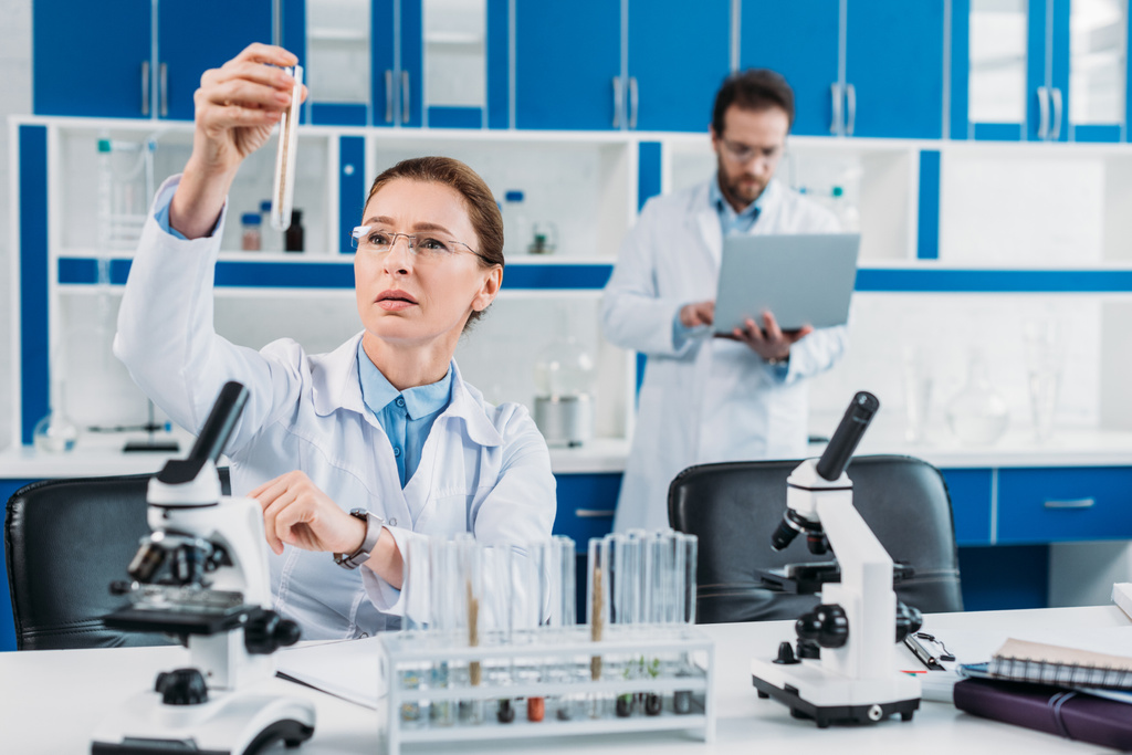 foco seletivo de cientista feminino olhando para tubo com reagente na mão com colega atrás no laboratório
 - Foto, Imagem