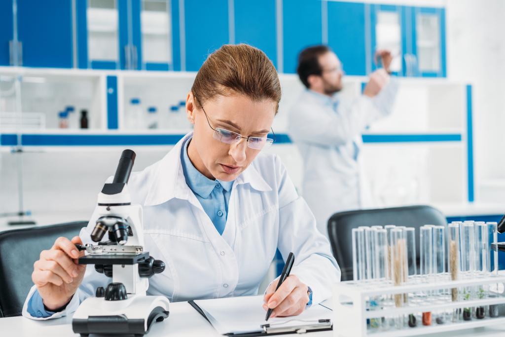 enfoque selectivo de la científica femenina haciendo notas en el bloc de notas en el lugar de trabajo con su colega detrás en el laboratorio
 - Foto, imagen