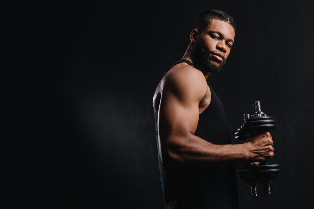 μυϊκή αφροαμερικάνος αθλητής κρατώντας αλτήρες και βλέπουν τα φωτογραφικών μηχανών που απομονώνονται σε μαύρο - Φωτογραφία, εικόνα
