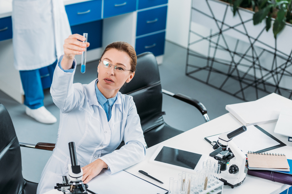 Naispuolisen tutkijan valikoiva keskittyminen putkeen reagenssin ollessa kädessä laboratoriossa takana olevan kollegan kanssa
 - Valokuva, kuva