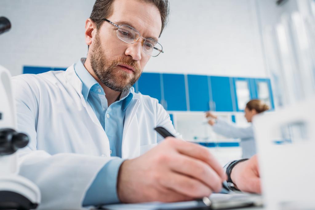 селективний фокус вченого в білому пальто і окулярах робить нотатки в блокноті на робочому місці в лабораторії
 - Фото, зображення