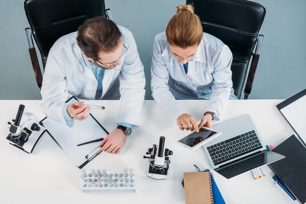 Κάτοψη των επιστημονικών ερευνητών σε λευκές στρώσεις χρησιμοποιώντας tablet μαζί στο χώρο εργασίας στο εργαστήριο - Φωτογραφία, εικόνα