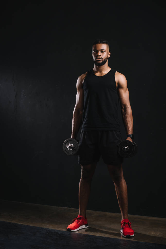 vista completa de joven muscular afroamericano deportista sosteniendo mancuernas en negro
 - Foto, Imagen