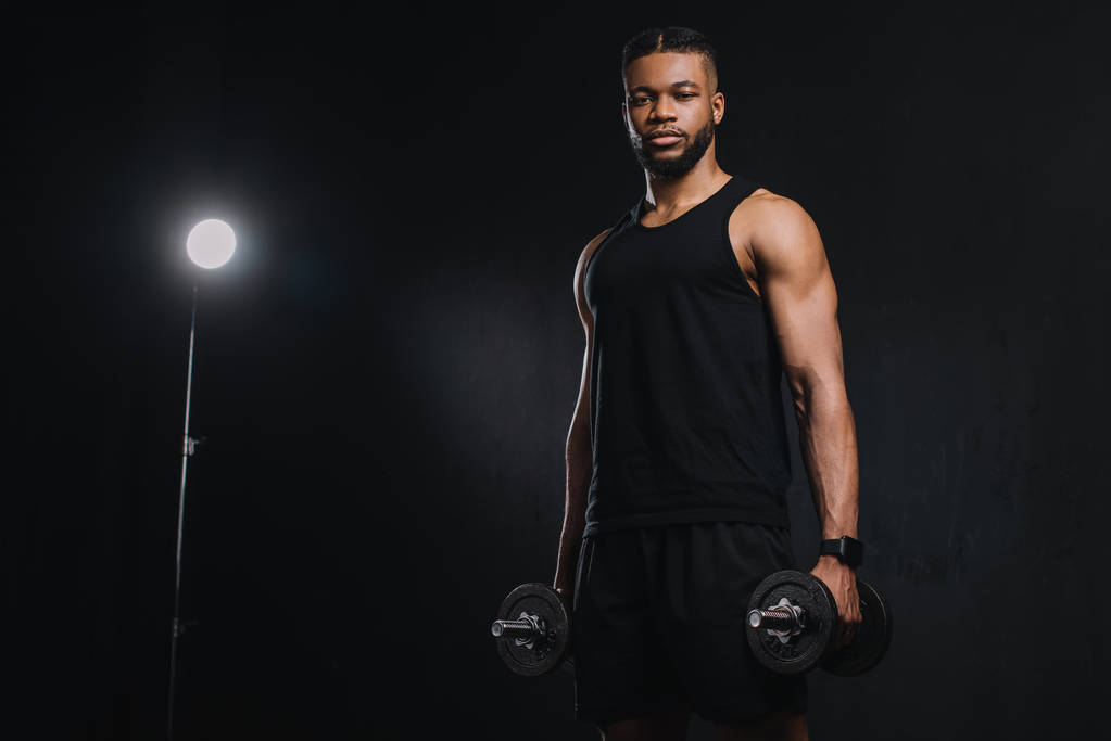 mladý americký sportovec drží činky a při pohledu na fotoaparát na černém - Fotografie, Obrázek
