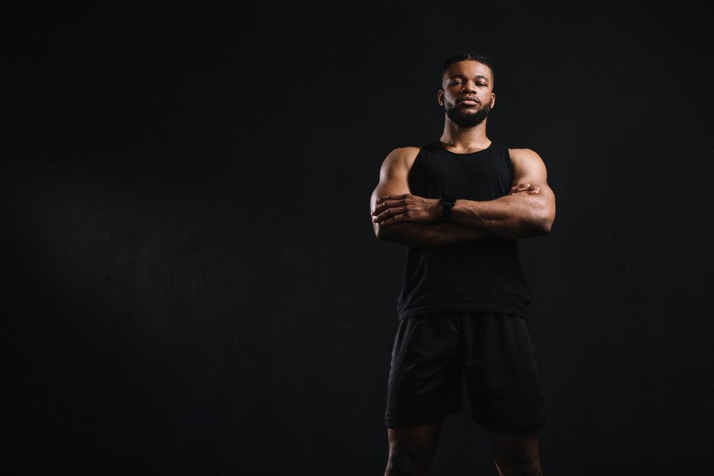 χαμηλή γωνία θέα αυτοπεποίθηση αφροαμερικάνος αθλητής στέκεται με σταυρωμένα τα χέρια και κοιτάζοντας φωτογραφική μηχανή που απομονώνονται σε μαύρο - Φωτογραφία, εικόνα