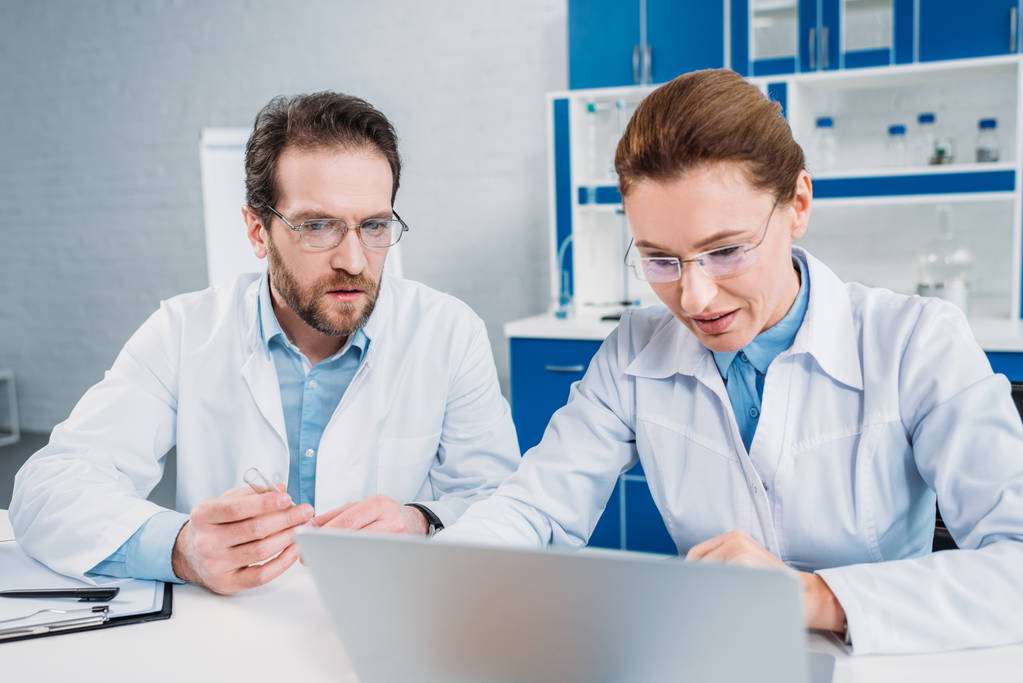 πορτρέτο του επιστήμονες σε ρόμπες και γυαλιά που εργάζονται για το laptop μαζί στο χώρο εργασίας στο εργαστήριο - Φωτογραφία, εικόνα