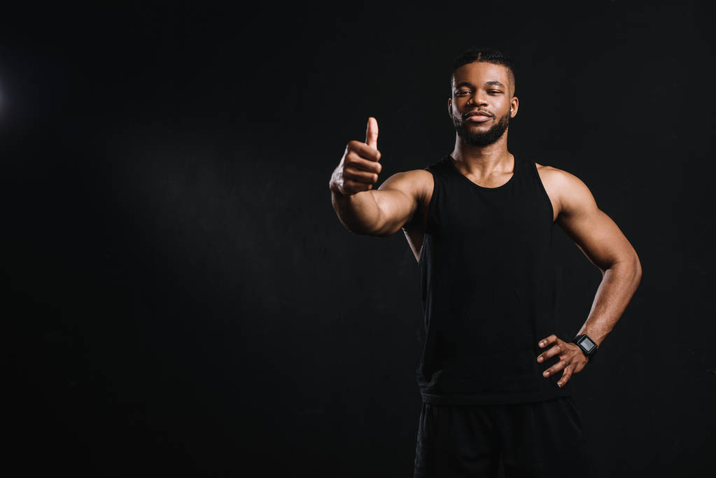 athletischer junger afrikanisch-amerikanischer Mann zeigt Daumen nach oben und schaut isoliert in die Kamera auf schwarz - Foto, Bild