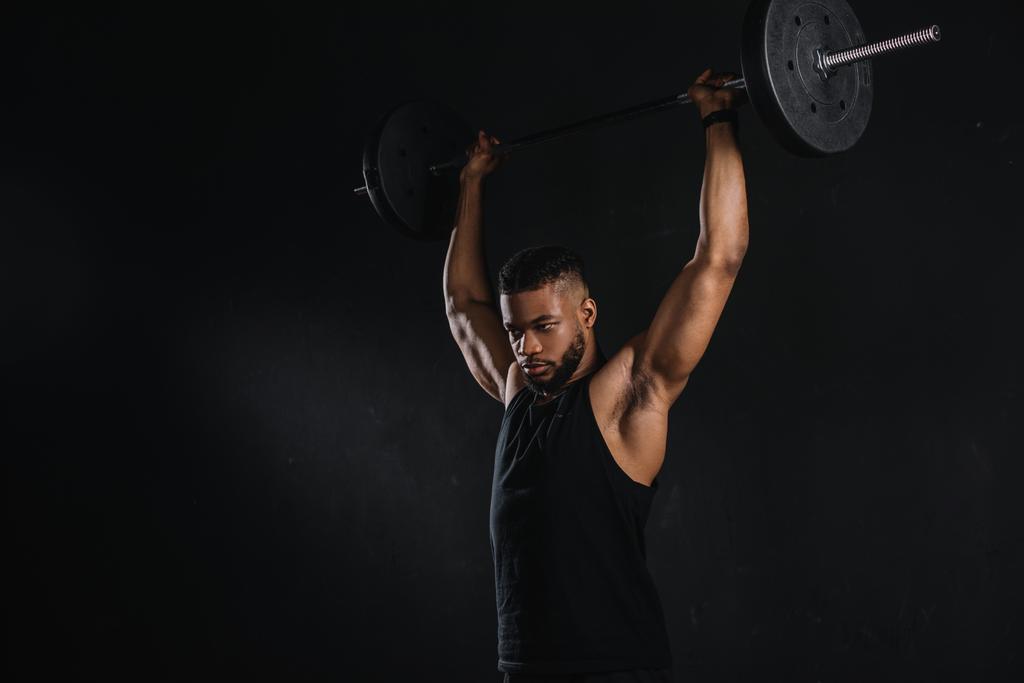 muscular young african american sportsman lifting barbell and looking away isolated on black - Photo, Image