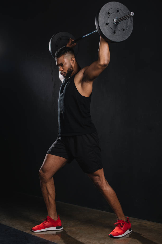 volle lengte weergave van gespierde jonge Afro-Amerikaanse sportman opheffing barbell op zwart  - Foto, afbeelding