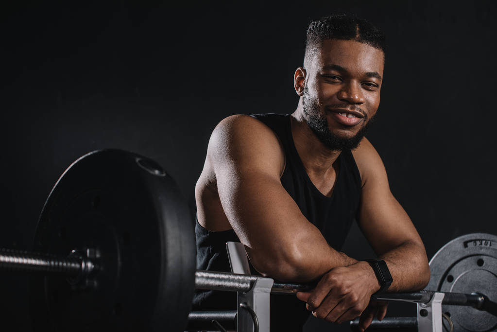 м'язистий молодий афроамериканський спортсмен, спираючись на барбелла і посміхаючись на камеру на чорному
 - Фото, зображення