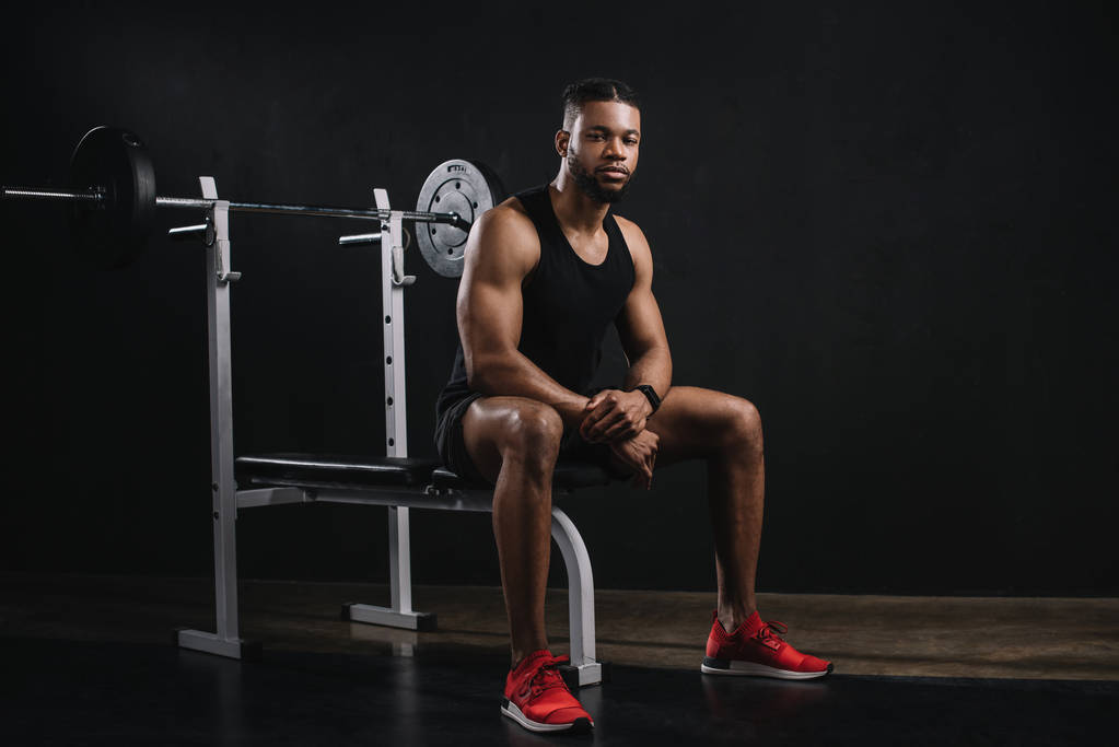 lihaksikas nuori afrikkalainen amerikkalainen mies urheiluvaatteissa istuu ja katselee kameraa kuntosalilla
 - Valokuva, kuva