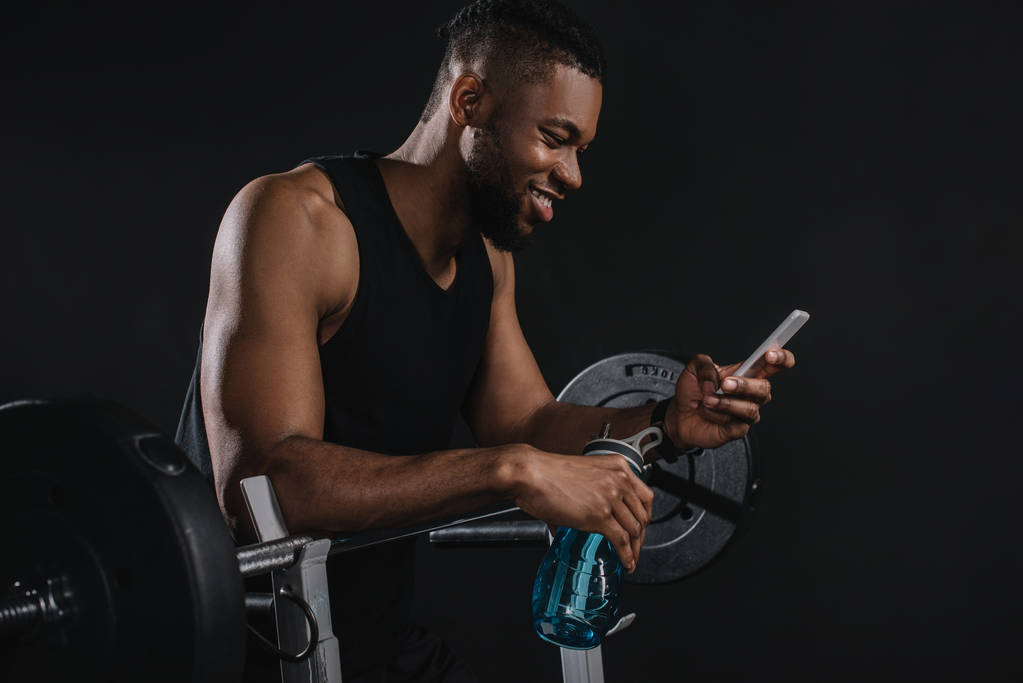 笑顔の若いアフリカ系アメリカ人のスポーツマンはスマート フォンを使用し、水のボトルを保持 - 写真・画像